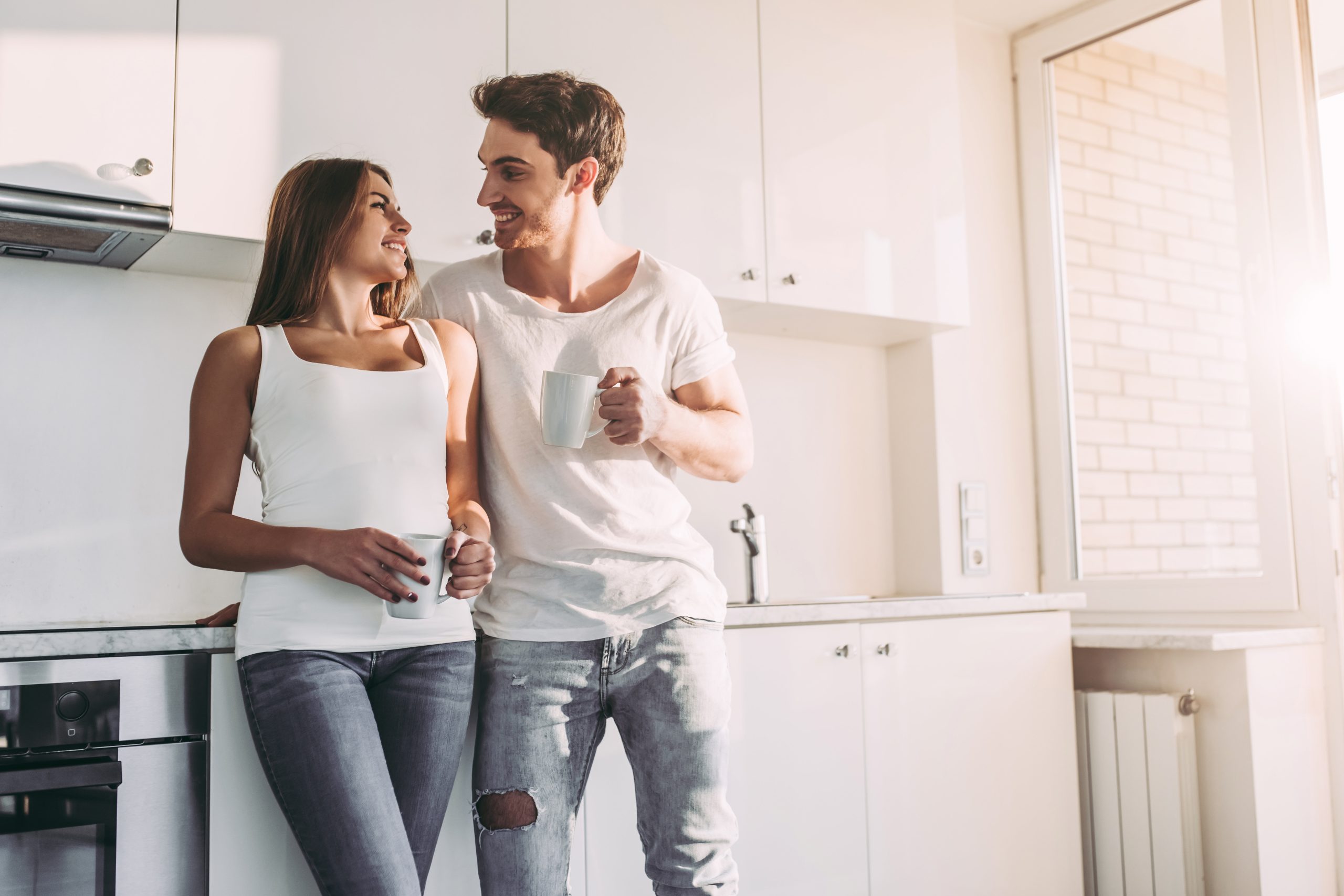 Romantic couple at home. Attractive young woman and handsome man are enjoying spending time together while standing on light modern kitchen with cup of coffee in hands.